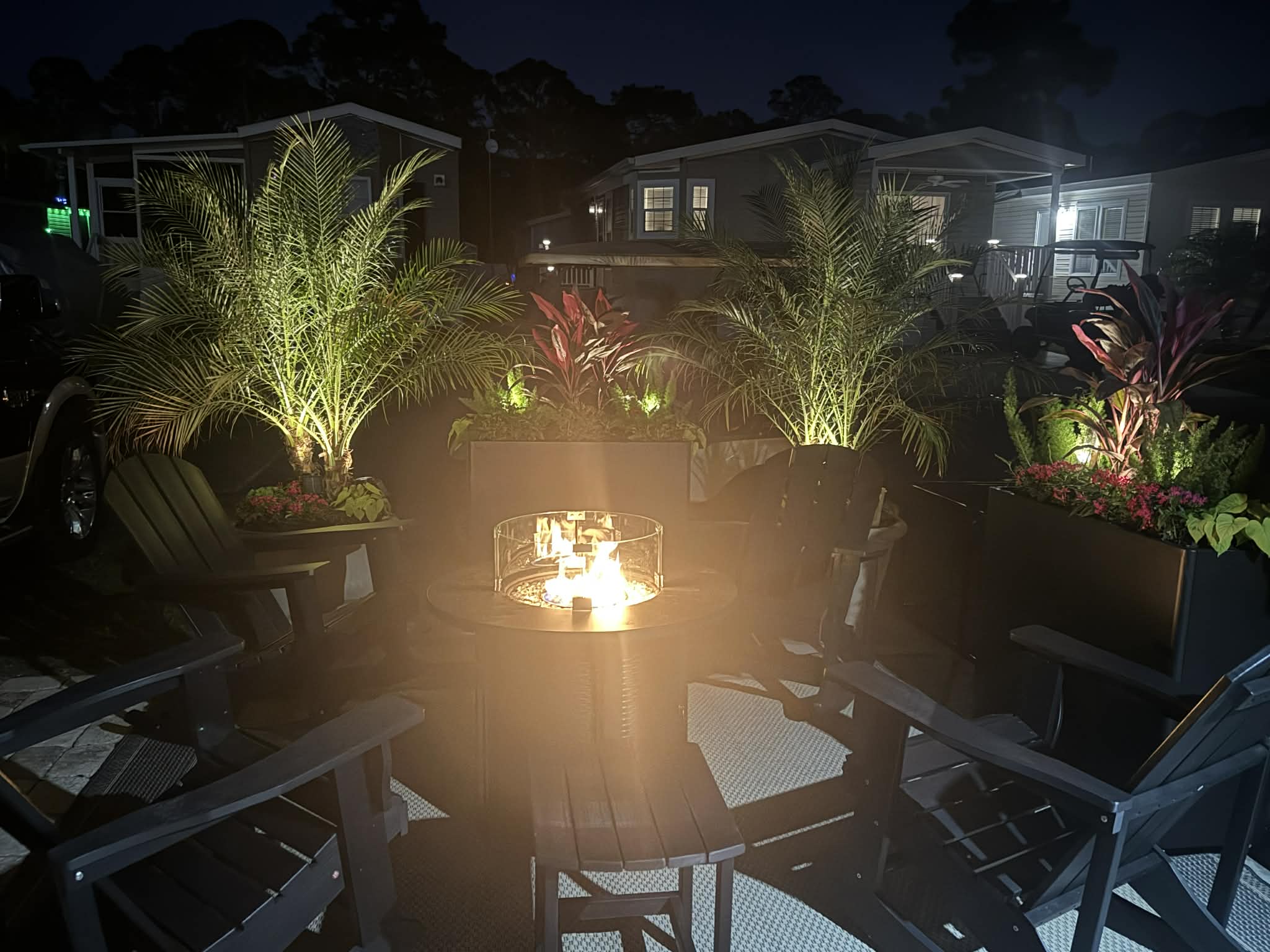 Outdoor fire table lit in the evening, with rocking chairs and illuminated tropical planters